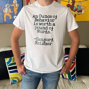 Può includere: Maglietta bianca con una citazione nera che recita "Un'oncia di comportamento vale una libbra di parole. -Sanford Meisner"