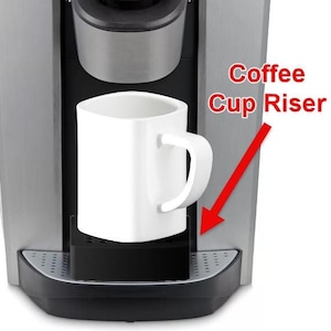 May include: A silver coffee maker with a removable cup riser. The coffee maker has a green coffee pod in the top and a white mug on the cup riser.