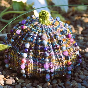 May include: A knitted pumpkin-shaped hat with a green top and decorated with purple, blue, and silver beads.