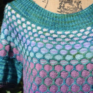 May include: A teal, purple, and green knitted sweater with a honeycomb pattern. The sweater has a round neckline and short sleeves.