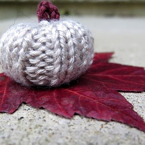 May include: A small knitted pumpkin in a light beige colour sits on a red maple leaf. The pumpkin has a burgundy stem.