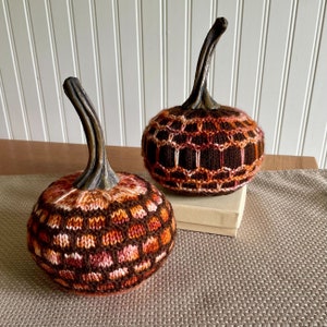 May include: Two knitted pumpkins in shades of brown, orange, and pink. The pumpkins have a geometric pattern and are sitting on a brown surface.