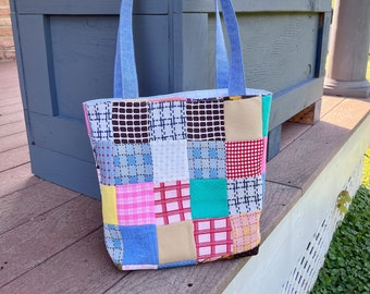 May include: A patchwork tote bag with blue handles. The bag is made of various fabrics in pink, red, yellow, blue, and green. The bag is sitting on a wooden surface.
