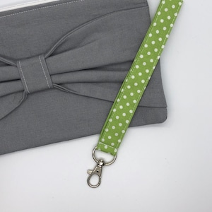 May include: A gray fabric pouch with a bow and a zipper closure. The pouch has a green and white polka dot wrist strap with a silver metal clasp.