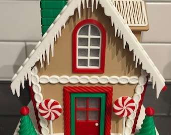Könnte beinhalten: Eine Lebkuchenhaus-Dekoration mit weißem Zuckerguss, roten Zuckerstangen und grünen Bäumen. Das Haus hat eine rote Tür, einen grünen Schornstein und ein weißes Dach mit Eiszapfen.
