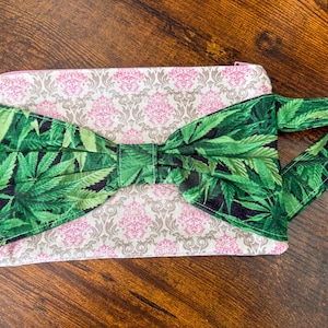 Może przedstawiać: Prostokątna kopertówka z zieloną kokardą i uchwytem w kształcie liścia konopi. Kopertówka ma różowo-szary wzór kwiatowy. Zamek błyskawiczny jest różowy. Na brązowej drewnianej powierzchni.