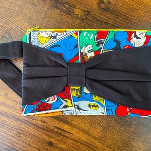 Puede incluir: Un bolso de mano rectangular con un lazo y asa negros. El bolso presenta un estampado de cómic con imágenes de superhéroes en colores primarios. La cremallera es amarilla.