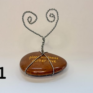 Puede incluir: Una piedra decorativa con las palabras "plant kindness gather love" en letras doradas. Un árbol de alambre plateado con una parte superior en forma de corazón está unido a la piedra. La piedra es marrón y tiene una superficie lisa.