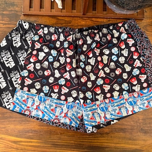 Puede incluir: Shorts negros, rojos y blancos con temática de Star Wars. Los shorts presentan varios patrones de personajes y logotipos de Star Wars, incluidos cascos de Darth Vader, cascos de Stormtrooper y el logotipo de Star Wars. Los shorts tienen una cintura elástica.