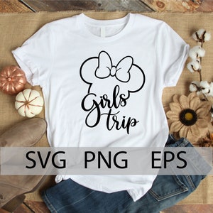 May include: White t-shirt with a black graphic of a Mickey Mouse head with a bow and the text "Girls Trip". The shirt is on a wooden surface with a pair of jeans and a brown shoe.