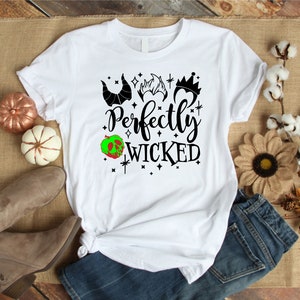 K&ouml;nnte beinhalten: Wei&szlig;es T-Shirt mit einem schwarzen grafischen Design, auf dem "Perfectly Wicked" mit einem gr&uuml;nen Apfel und schwarzen Silhouetten von Kronen und H&ouml;rnern steht.