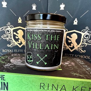 May include: A glass jar with a black lid and a label that reads "Kiss the Villain." The label features skulls, a cat silhouette, and crossed arrows. The jar sits on top of two books with green and black covers.