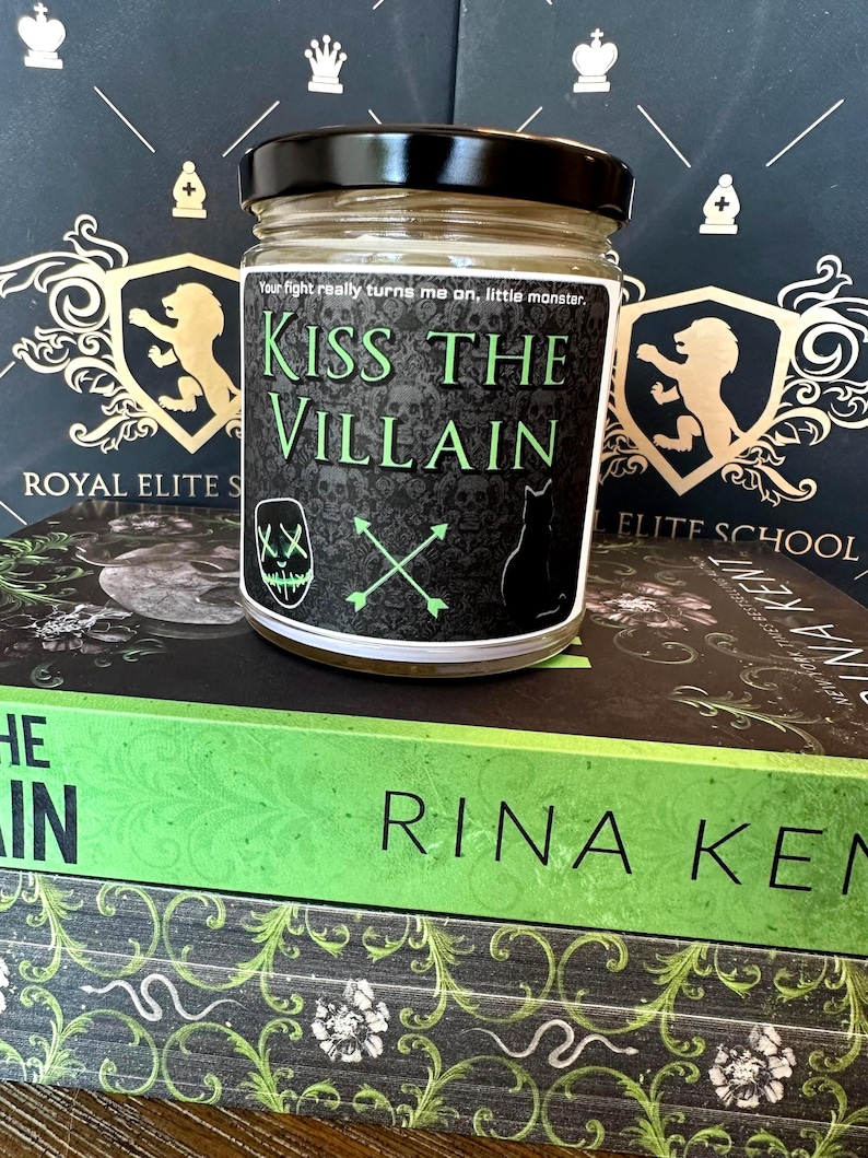 May include: A glass jar with a black lid and a label that reads "Kiss the Villain." The label features skulls, a cat silhouette, and crossed arrows. The jar sits on top of two books with green and black covers.