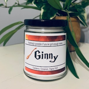 May include: A glass candle jar with a black lid. The label is red and white with the text "Anything's possible if you've got enough nerve. Ginny Cranberry. Cinnamon. Pygmy Puff."
