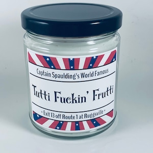 Puede incluir: Una vela de vidrio en un frasco con tapa negra. La etiqueta es roja, blanca y azul con estrellas y el texto "Captain Spaulding's World Famous Tutti Fuckin' Frutti * Exit 13 off Route 1 at Ruggsville *"