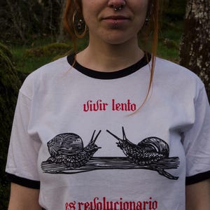 May include: White t-shirt with black trim featuring two snail illustrations and the text "vivir lento es revolucionario" in red. The shirt has a crew neck and short sleeves.