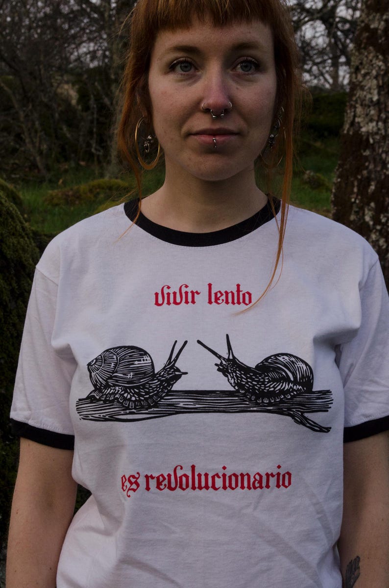 May include: White t-shirt with black trim featuring two snail illustrations and the text "vivir lento es revolucionario" in red. The shirt has a crew neck and short sleeves.