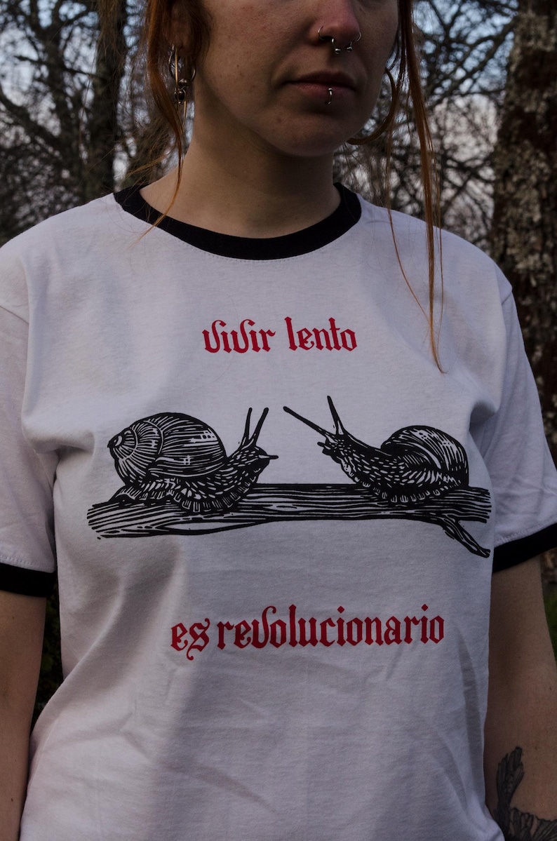 May include: White t-shirt with black trim featuring a black and white illustration of two snails facing each other on a branch. The red text above reads "Vivir lento" and below "es revolucionario".