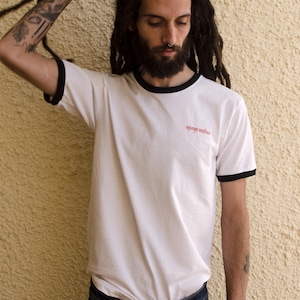 May include: A white t-shirt with black trim around the neck and sleeves. The shirt has the words "apoyo mutuo" printed in red on the front.