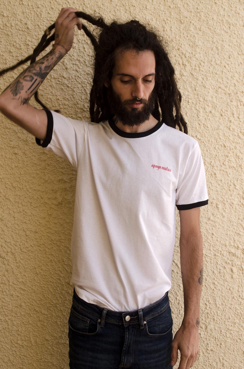 May include: A white t-shirt with black trim around the neck and sleeves. The shirt has the words "apoyo mutuo" printed in red on the front.