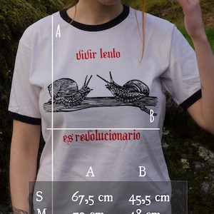 May include: White t-shirt with black trim featuring the Spanish text "Vivir lento es revolucionario" and a graphic of two snails. The shirt's measurements are: S - 67.5 cm x 45.5 cm, M - 70 cm x 48 cm, L - 72.5 cm x 53 cm, XL - 75 cm x 58.5 cm.