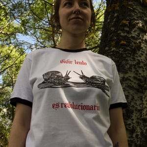 May include: White t-shirt with black trim featuring a graphic of two snails and the text "vivir lento es revolucionario" in red. The shirt is worn by a person outdoors, with trees in the background.