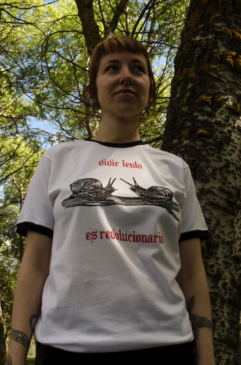 May include: White t-shirt with black trim featuring a graphic of two snails and the text "vivir lento es revolucionario" in red. The shirt is worn by a person outdoors, with trees in the background.
