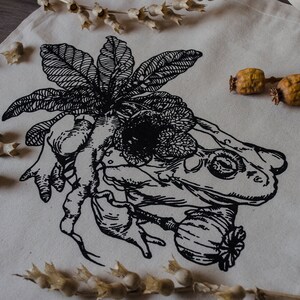 Op de afbeelding: Een zwart-witte tote bag met een grafische print van een kikker, een bloem en een plant. Het ontwerp doet denken aan een vintage botanische illustratie.