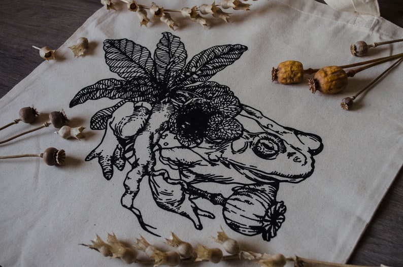 Op de afbeelding: Een zwart-witte tote bag met een grafische print van een kikker, een bloem en een plant. Het ontwerp doet denken aan een vintage botanische illustratie.