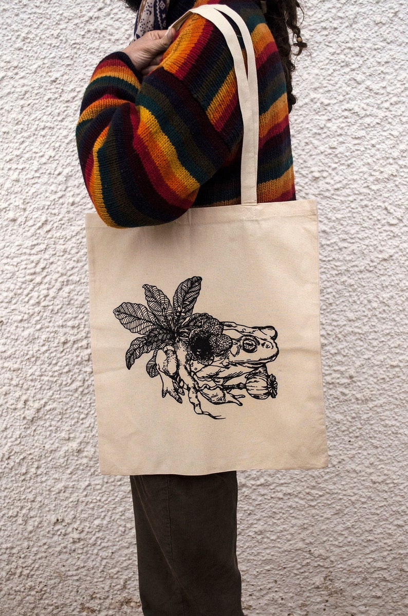 Op de afbeelding: Een beige tote bag met een zwart-witte illustratie van een kikker die op een blad zit. De kikker heeft een groot oog en is omringd door andere planten.