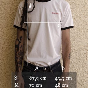 May include: White t-shirt with black trim around the neck and sleeves. The shirt has the text "apoyo mutuas" printed on the front. Size chart with measurements in centimeters for different sizes: S, M, L, XL.