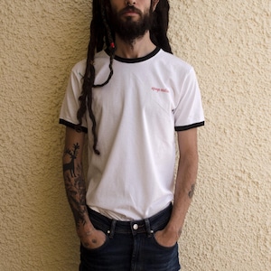 May include: A man with long dreadlocks is wearing a white t-shirt with black trim and the text "apoyo mutuo" in red. He is wearing blue jeans and has tattoos on his arms.