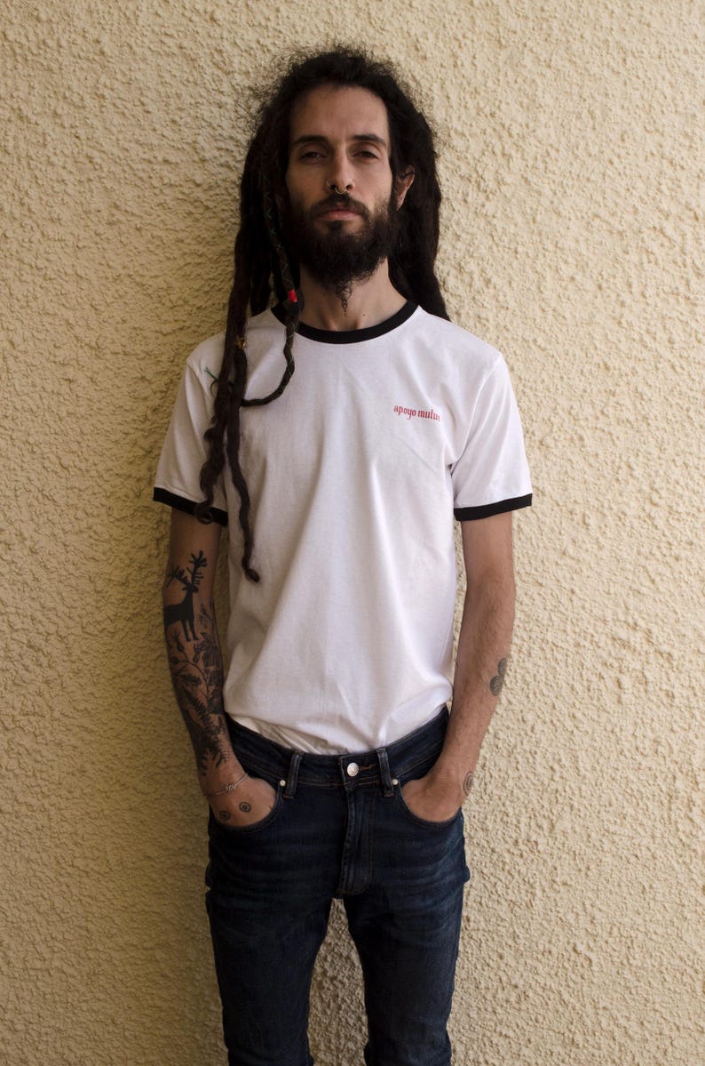May include: A man with long dreadlocks is wearing a white t-shirt with black trim and the text "apoyo mutuo" in red. He is wearing blue jeans and has tattoos on his arms.