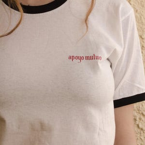 May include: White t-shirt with a black ringer and the text "apoyo mutuo" in red.