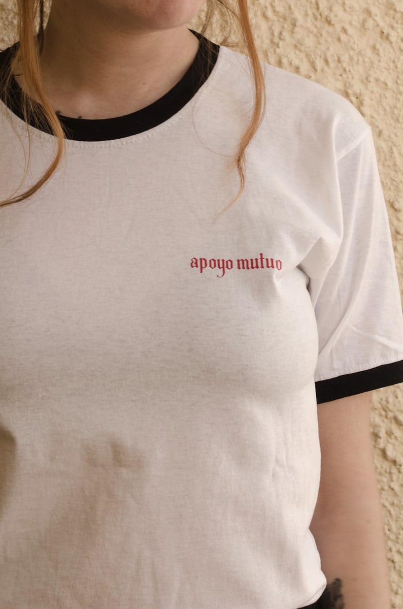 May include: White t-shirt with a black ringer and the text "apoyo mutuo" in red.