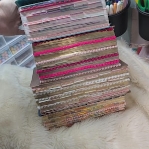 May include: A stack of four books with gold-gilded page edges, each featuring decorative pom-pom trim in shades of pink, white, and orange attached to the page edges, along with numerous colorful adhesive index tabs protruding from the sides.