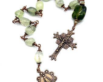 Könnte beinhalten: Ein Rosenkranz mit hellgrünen, ovalen Perlen und einem bronzenen Kruzifix mit einer detaillierten Darstellung Jesu. Der Rosenkranz enthält auch ein Bronze-Medaillon mit einer religiösen Figur.
