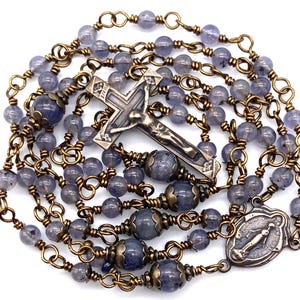 May include: A rosary featuring translucent light purple beads linked by antique bronze wire. The piece includes a central bronze crucifix with the letters INRI at the top and a rectangular bronze medal depicting a figure in relief at the bottom.