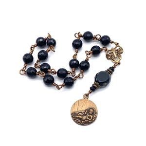 May include: A rosary featuring faceted black beads linked by copper-toned wire. The piece includes a central connector with a small cross and a round bronze-toned medal depicting a figure lying down, finished with a larger black bead above it.