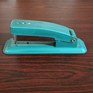 May include: A vintage teal Swingline CUB stapler. The stapler has the text "Swingline CUB" and "LIFT TO LOAD" printed on it. The stapler is made of metal and has a rectangular base.