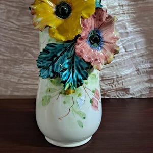 May include: A white ceramic vase with a floral design. The vase has three dimensional flowers, including a yellow flower, a pink flower, and green leaves.