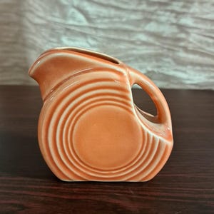 May include: A small, orange ceramic pitcher with a handle and a spout. The pitcher has a circular design on the front and is set on a dark wooden surface. The background is a light, textured fabric.