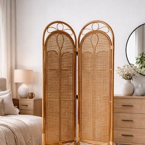 Op de afbeelding: Een vrijstaand kamerscherm met twee panelen van natuurlijk rotan. De scheidingswand heeft een geweven rietontwerp in een lichtbruin houten frame. De bovenkant van elk paneel heeft een boogontwerp met decoratieve details.