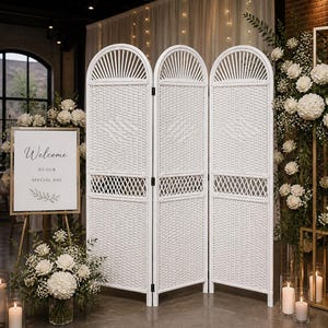 Puede incluir: Un biombo blanco tejido con una parte superior arqueada, preparado para una boda o evento especial. Un letrero que dice "Bienvenido a nuestro d&iacute;a especial" est&aacute; frente al biombo. Flores blancas y velas tambi&eacute;n est&aacute;n en la escena.