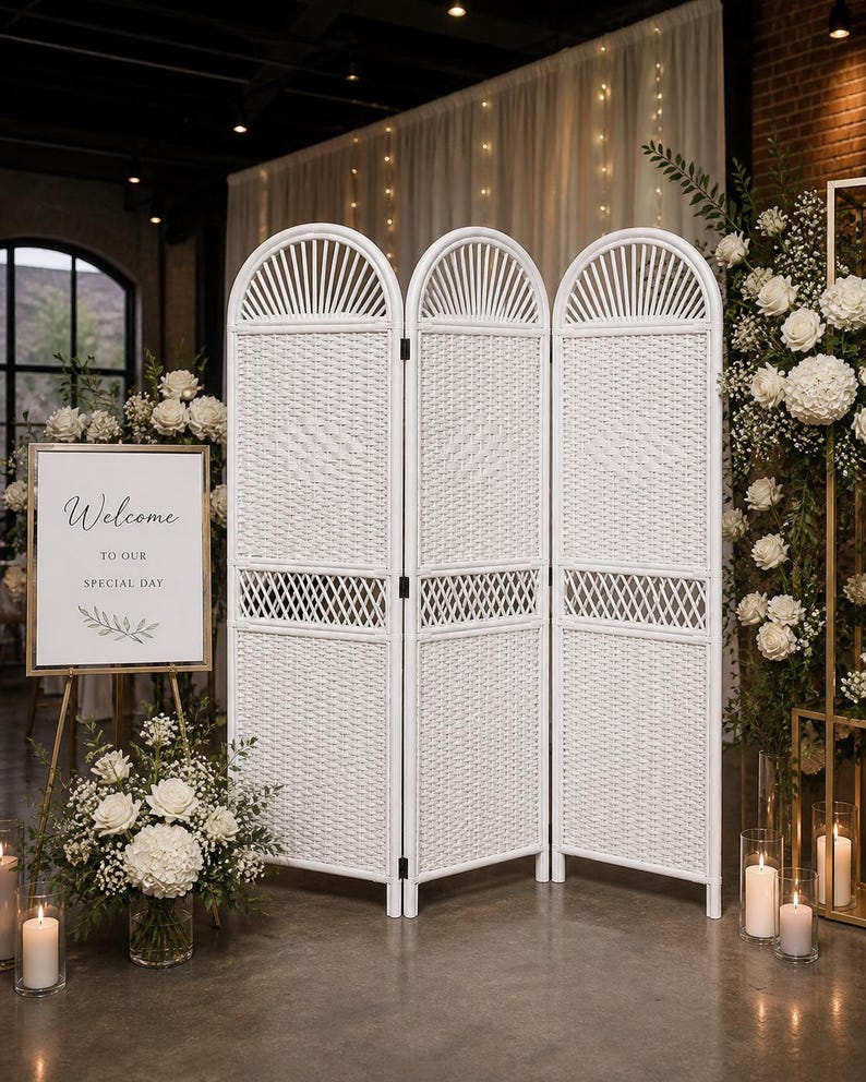 Puede incluir: Un biombo blanco tejido con una parte superior arqueada, preparado para una boda o evento especial. Un letrero que dice "Bienvenido a nuestro d&iacute;a especial" est&aacute; frente al biombo. Flores blancas y velas tambi&eacute;n est&aacute;n en la escena.