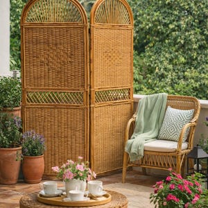 Puede incluir: Un biombo de mimbre natural con la parte superior arqueada se encuentra en un patio, junto a una silla de mimbre con una manta verde y un coj&iacute;n estampado. Una mesa de centro redonda de mimbre sostiene una bandeja con tazas y un jarr&oacute;n con flores rosas. Plantas en macetas rodean el patio.