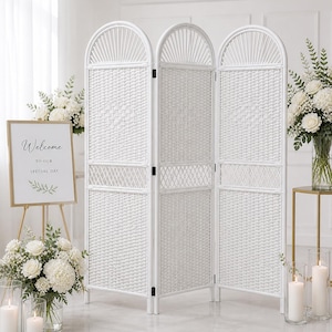 Puede incluir: Un biombo blanco de tres paneles tejido con arcos, ideal para eventos. Un letrero con marco dorado con la palabra "Bienvenida" est&aacute; cerca. Arreglos florales blancos y velas en portavelas de vidrio completan la escena.
