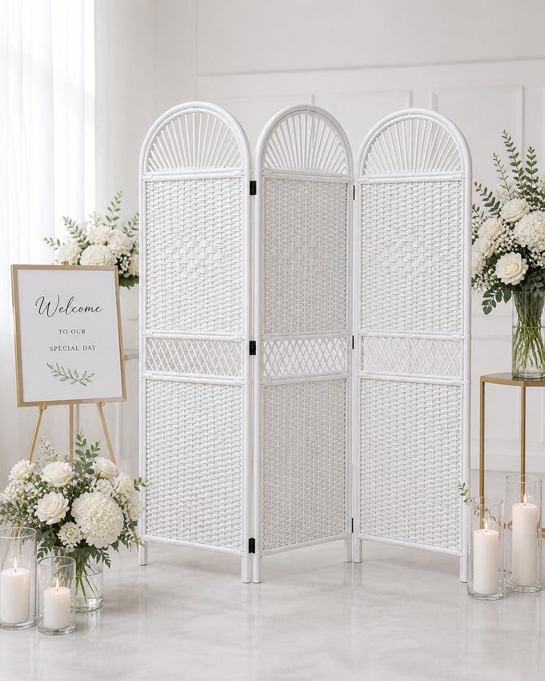 Puede incluir: Un biombo blanco de tres paneles tejido con arcos, ideal para eventos. Un letrero con marco dorado con la palabra "Bienvenida" est&aacute; cerca. Arreglos florales blancos y velas en portavelas de vidrio completan la escena.