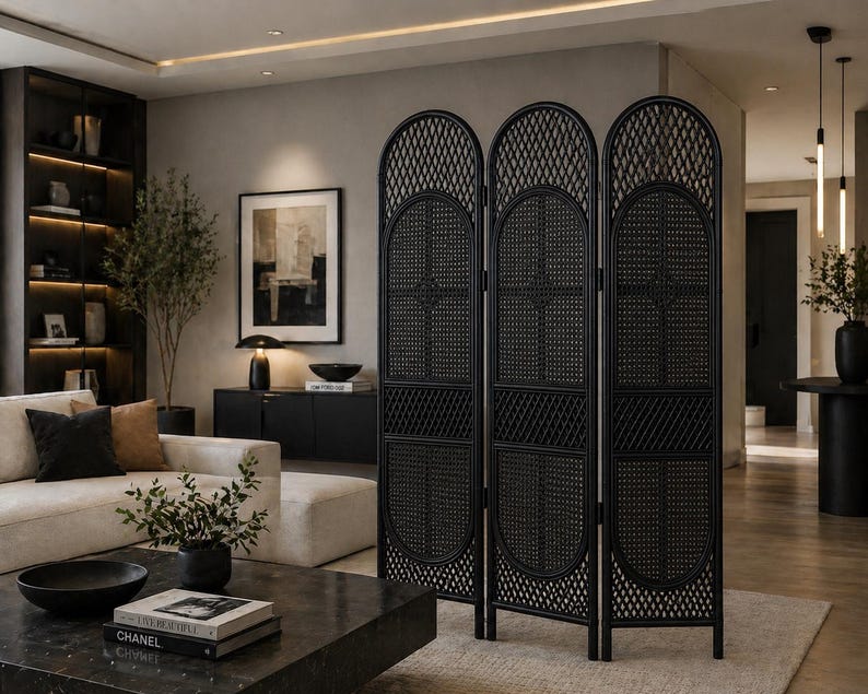 May include: A black, three-panel room divider with an arched top and woven rattan design. The divider stands on a light-colored rug in a modern living room setting. The room features a sofa, artwork, and decorative plants.