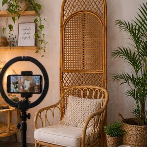 May include: A cozy interior scene featuring a rattan chair with a cream cushion and a macrame pillow. A tall, arched rattan screen stands behind the chair. A ring light with a phone is in the foreground. Plants and a mug with text add to the warm, inviting atmosphere.